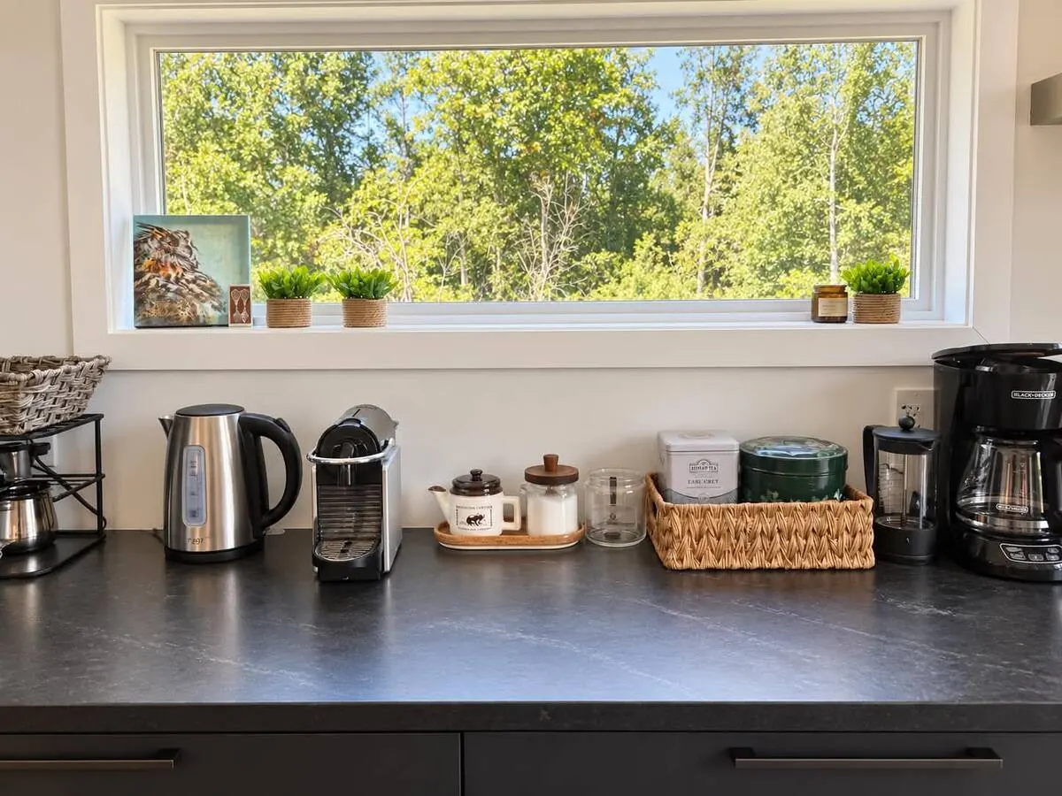 A coffee and tea station on a dark counter under a window looking into the trees