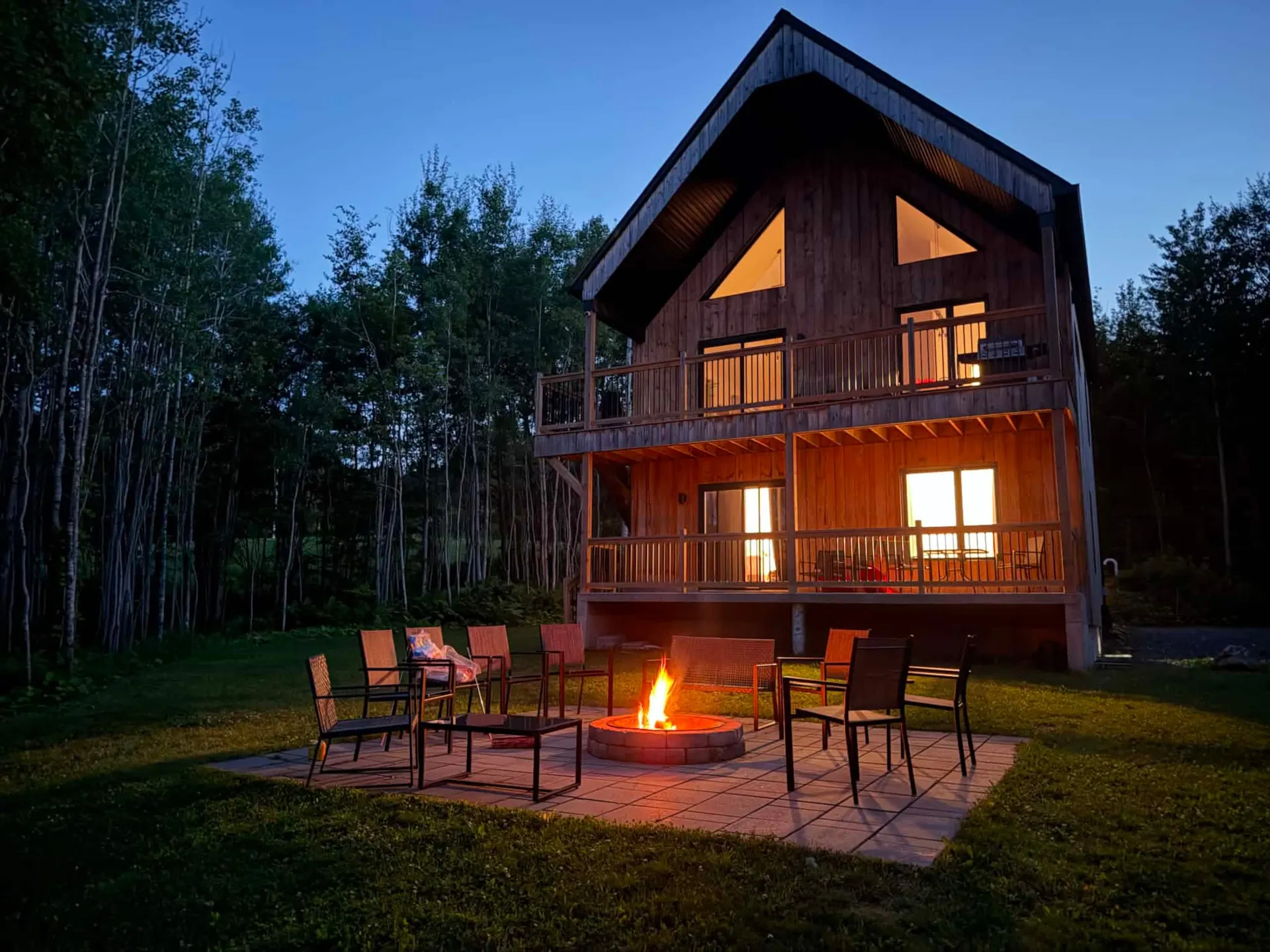 La Villa Foresta at dusk: a tall wooden chalet with glowing windows, chairs set around a lit fire pit on a stone patio